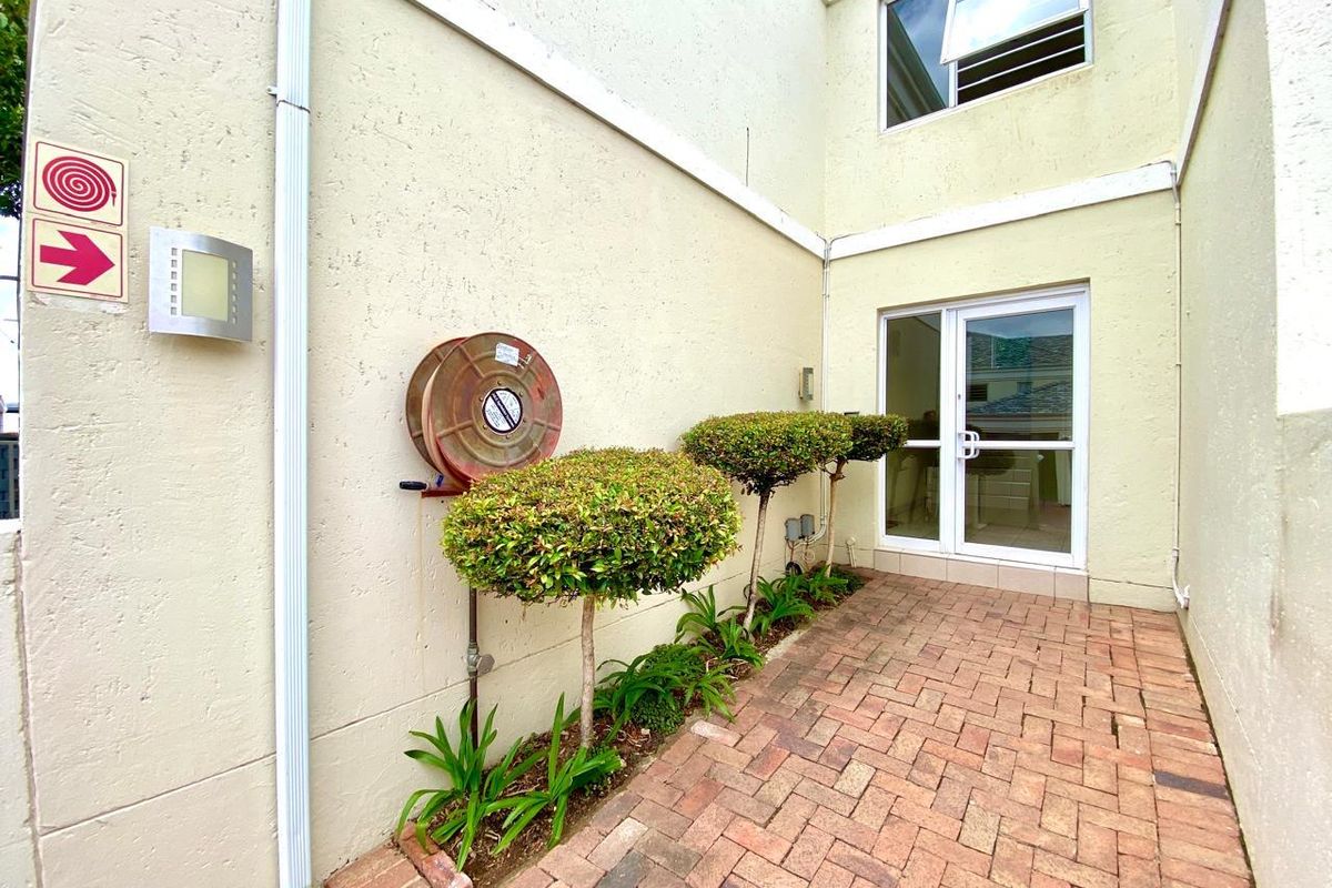 A paved rear courtyard area featuring a fire hose reel, landscaped shrubs, and access to the apartment via a glass sliding door.