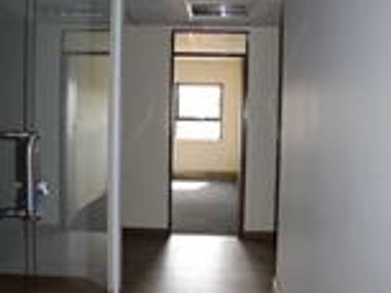 A functional office hallway featuring laminate flooring, a glass partition wall, and recessed ceiling lighting leading into an adjacent office space.