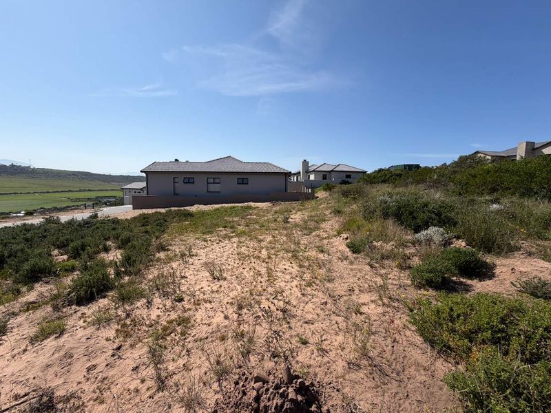A view of a vacant residential plot featuring sandy terrain and native vegetation, with neighboring properties visible in the background.