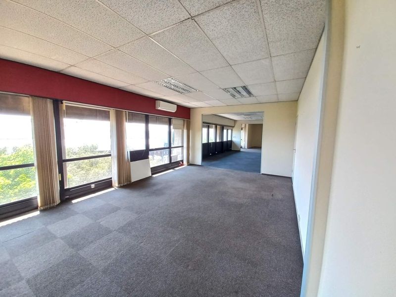 Spacious open-plan office area featuring large windows, carpeted flooring, suspended ceiling tiles, and a wall-mounted air conditioning unit.