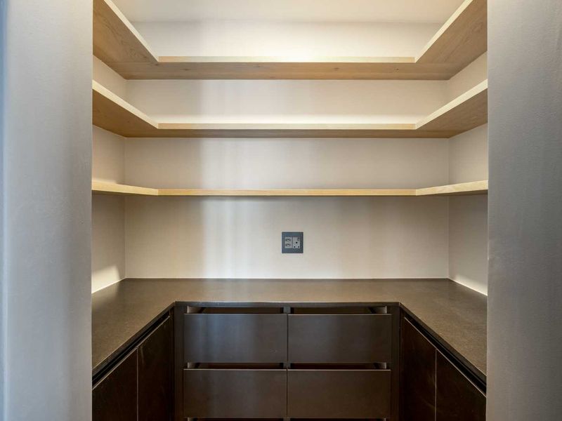 A modern, well-appointed scullery or pantry featuring sleek dark cabinetry, stone countertops, and ample floating timber shelving for organized storage.