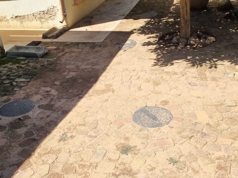 An outdoor paved area featuring cobblestone surfacing and visible drainage covers, showing signs of wear and weathering on the adjacent building structure.