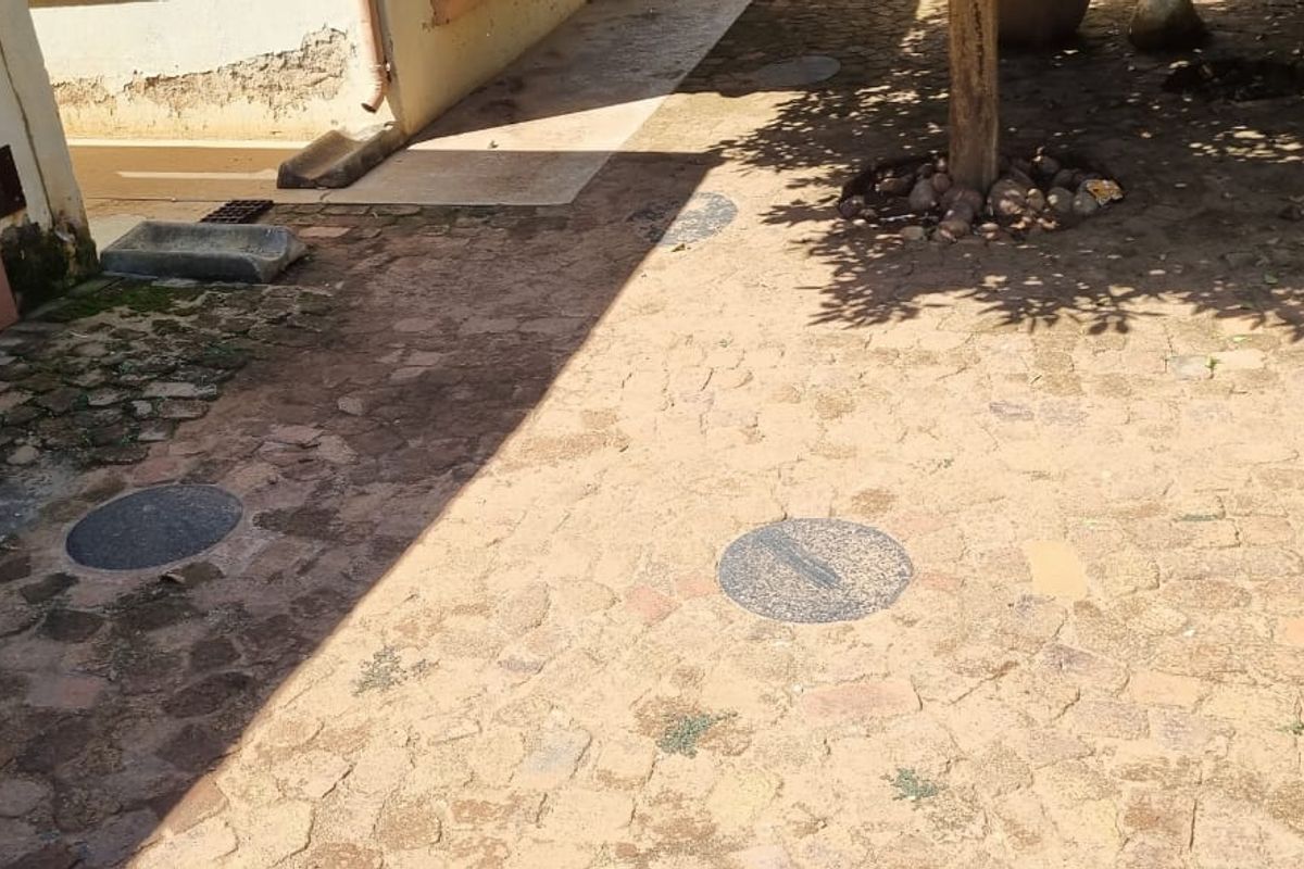 An outdoor paved area featuring cobblestone surfacing and visible drainage covers, showing signs of wear and weathering on the adjacent building structure.