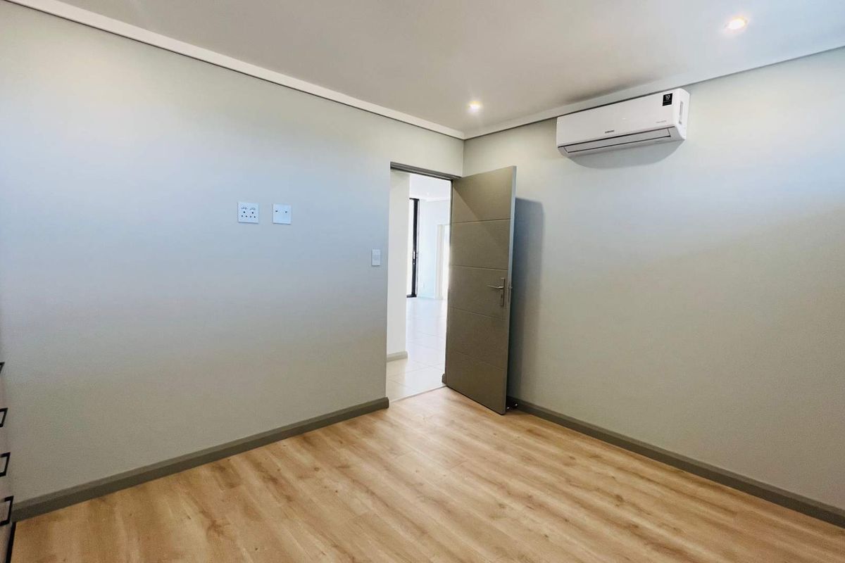 A bright, modern bedroom featuring light wood-look flooring, neutral walls, a wall-mounted air conditioning unit, and a built-in wardrobe.