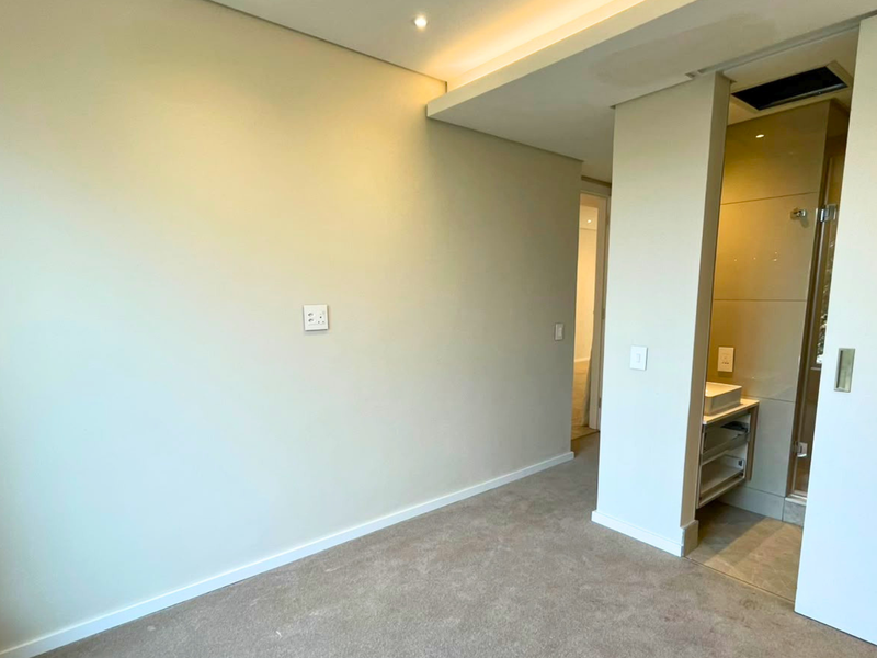 A clean, empty bedroom featuring neutral carpeting, recessed ceiling lighting, and a modern en-suite bathroom with a vanity basin and shower enclosure.