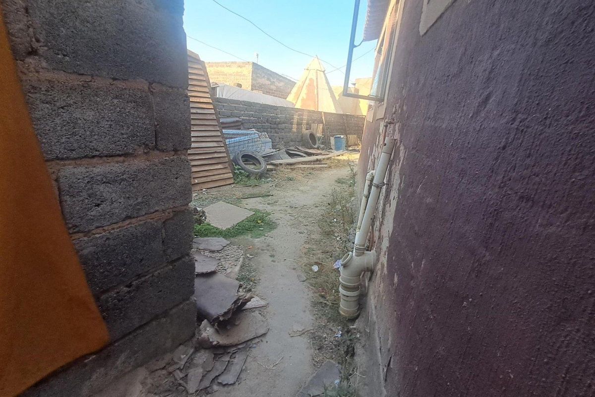 A narrow side passage between two masonry structures featuring exposed plumbing pipes and an unpaved ground surface requiring maintenance and clearing.