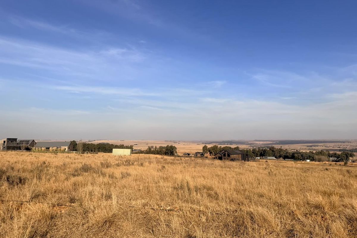 A wide-angle view of expansive, undeveloped residential land featuring dry grassland with distant neighboring properties under a clear blue sky.