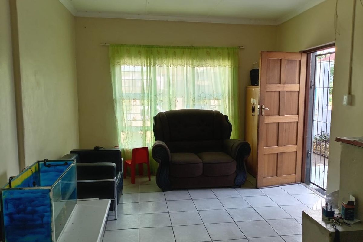 A living room area featuring tiled flooring, a wooden entrance door, and a window with sheer curtains.