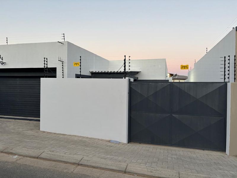 Modern front facade featuring a clean white perimeter wall, secure electric fencing, a dark metal driveway gate, and an automated garage door.