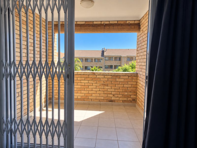 A private balcony featuring exposed brick walls, tiled flooring, and a security gate, offering an open-air space with views of the surrounding complex.