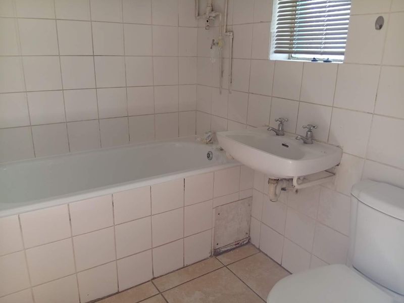 A dated bathroom featuring a bathtub, wall-mounted basin, and toilet, with exposed plumbing and tiled finishes requiring modernization and maintenance.