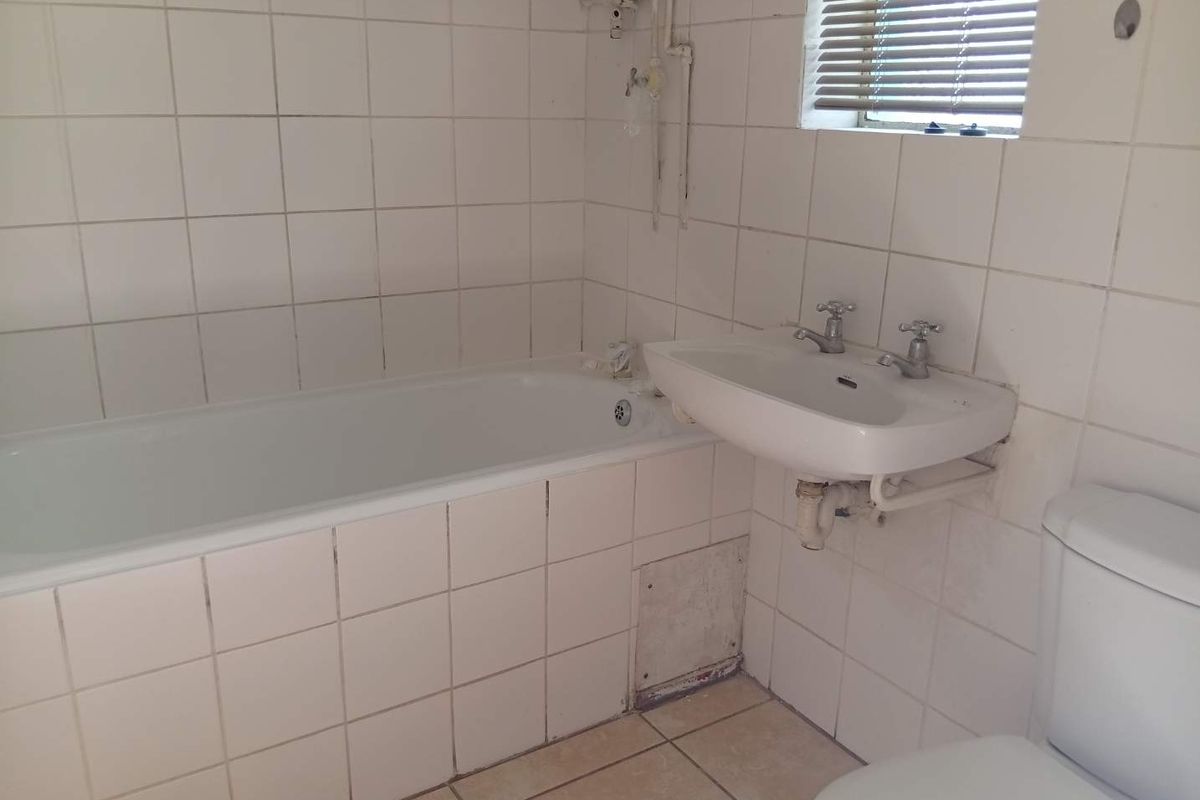 A dated bathroom featuring a bathtub, wall-mounted basin, and toilet, with exposed plumbing and tiled finishes requiring modernization and maintenance.
