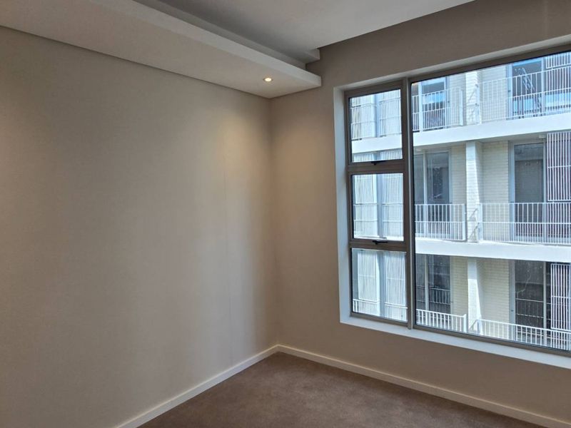 A bright, empty bedroom featuring neutral walls, carpeted flooring, recessed ceiling lighting, and a large window overlooking the apartment complex.