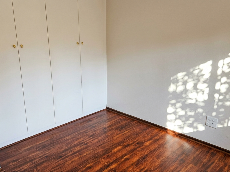 A bright, empty bedroom featuring dark laminate flooring and white built-in cupboards, offering a clean and neutral space ready for personalization.