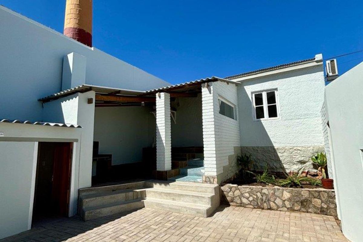 A paved rear courtyard featuring a covered patio area, stone-clad garden bed, and a prominent brick chimney stack.