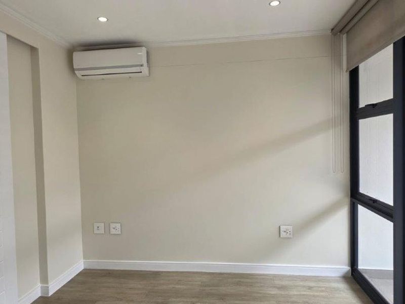 A bright, empty living area featuring modern laminate flooring, recessed lighting, a wall-mounted air conditioning unit, and large glass sliding doors with roller blinds.