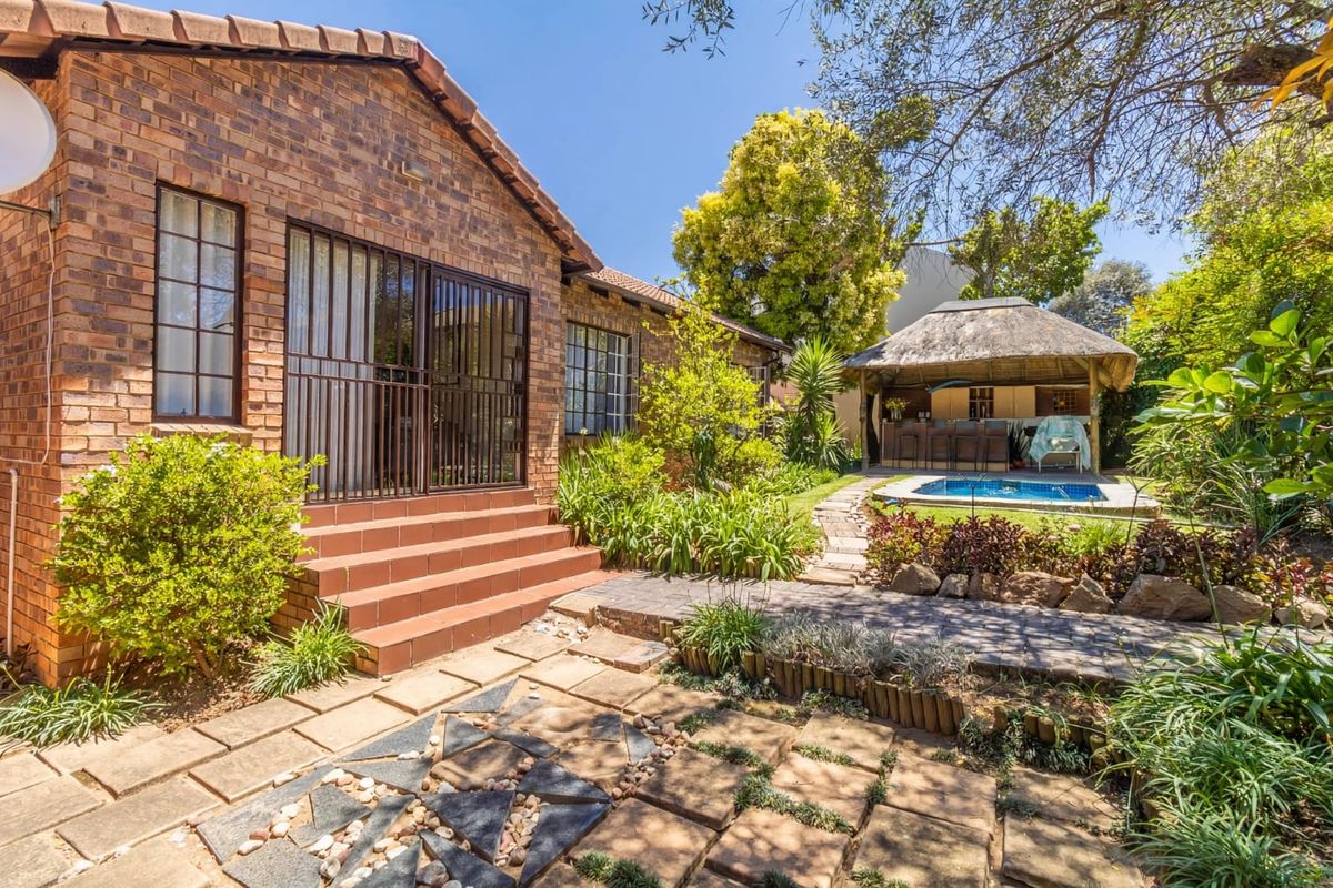 Rear view of the townhouse featuring a brick facade, paved patio area, swimming pool, and a thatched lapa set within a landscaped garden.
