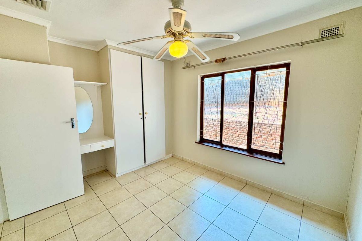 A vacant bedroom featuring tiled flooring, built-in cupboards, a built-in vanity, and a ceiling fan, with security bars installed on the window.