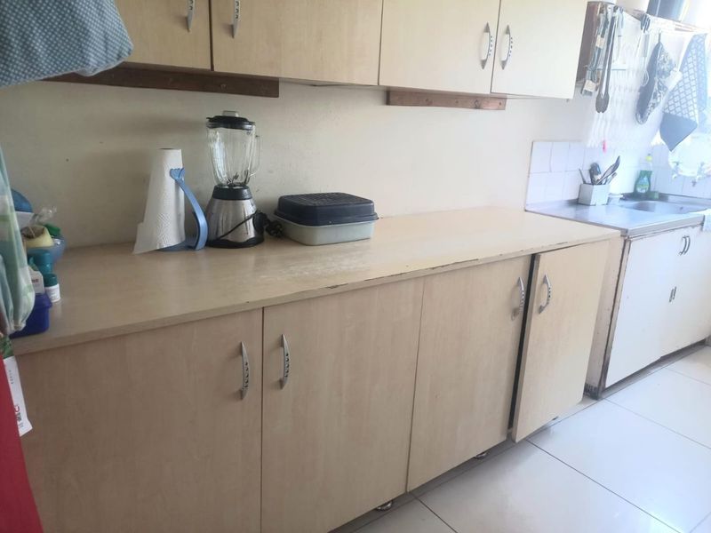 A functional kitchen area featuring light-toned laminate cabinetry, a stainless steel sink, and tiled splashback, showing signs of wear on the countertop surfaces.