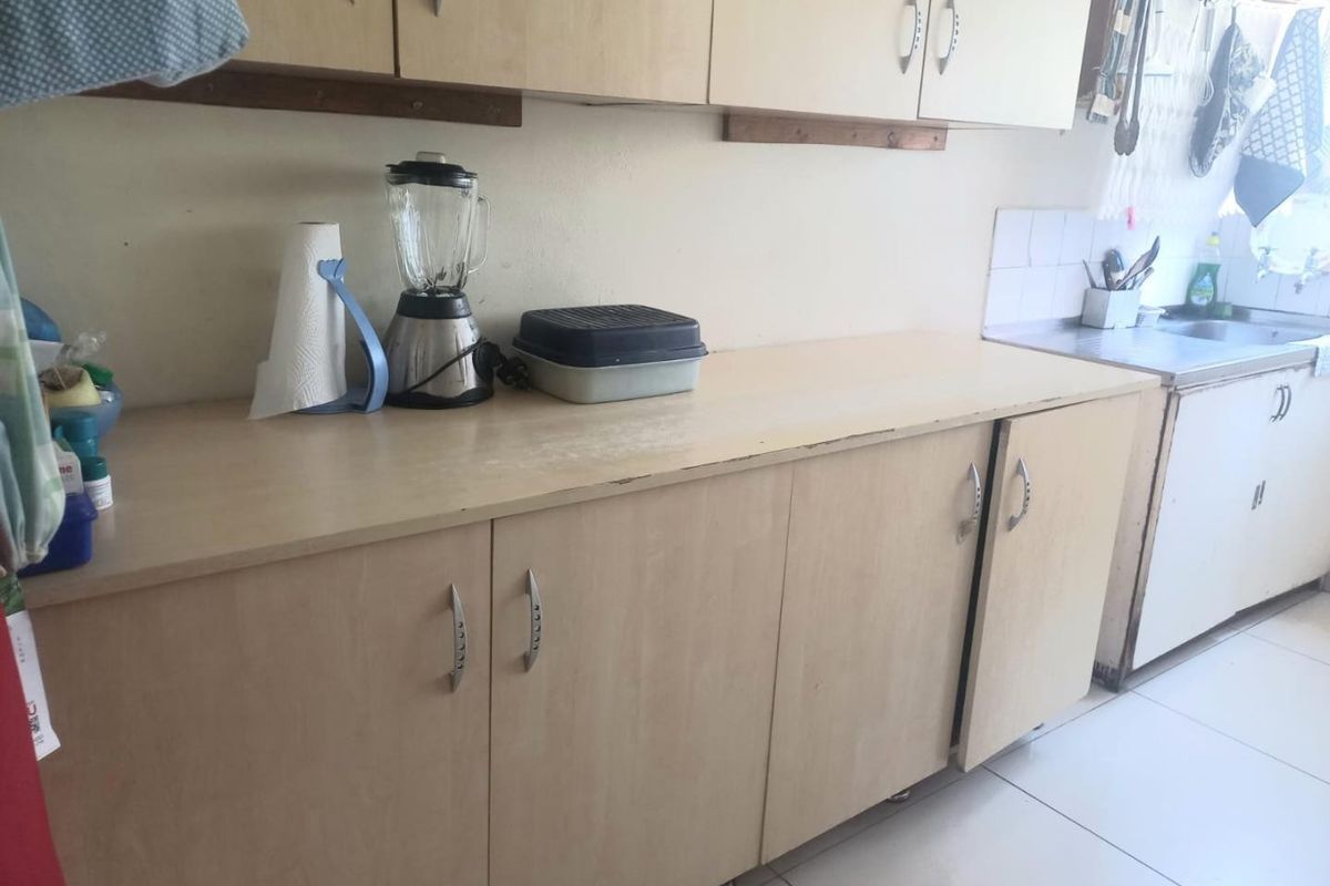 A functional kitchen area featuring light-toned laminate cabinetry, a stainless steel sink, and tiled splashback, showing signs of wear on the countertop surfaces.