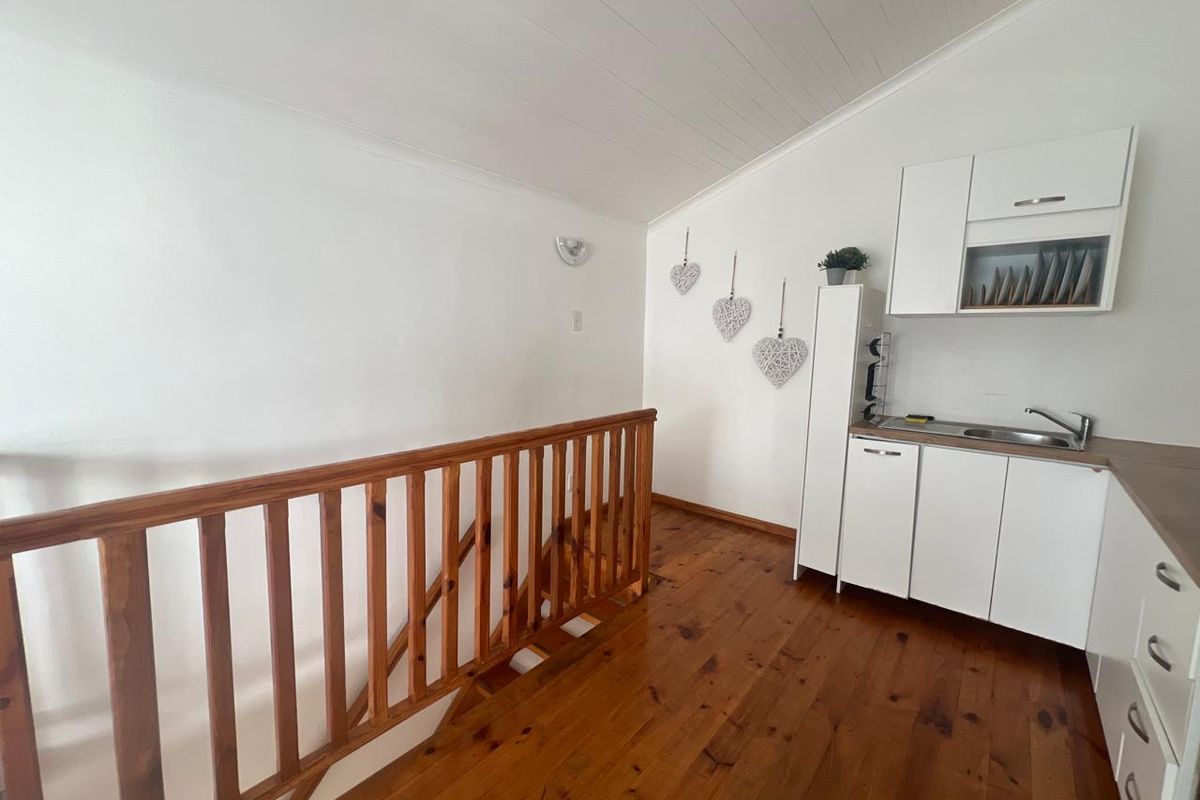A compact kitchen area featuring white cabinetry, a stainless steel sink, hardwood flooring, and a wooden staircase railing under a sloped ceiling.