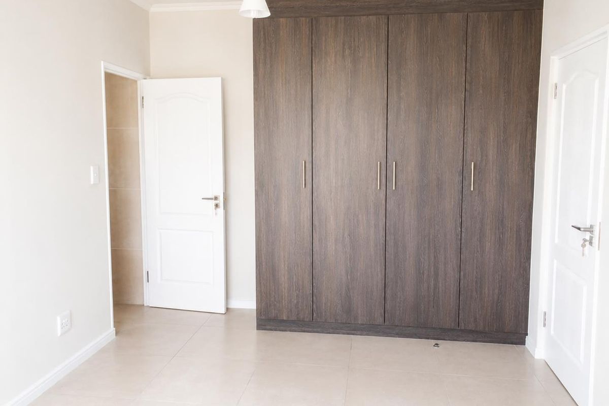 A clean, empty bedroom featuring modern dark wood built-in cupboards, neutral tiled flooring, and crisp white walls.