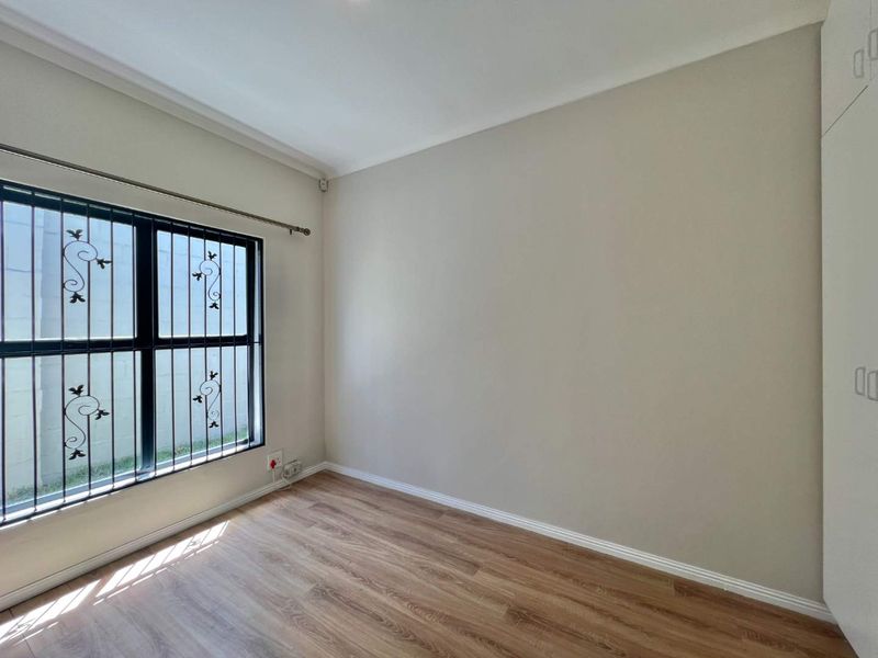 A bright, empty bedroom featuring modern laminate flooring, neutral walls, built-in cupboards, and a window fitted with security bars.