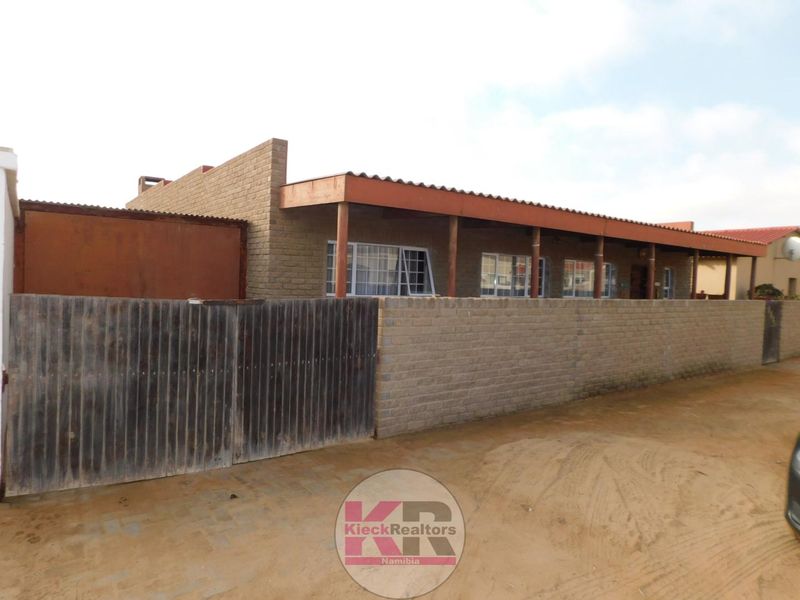 Front exterior view of a single-story house featuring a brick boundary wall, gated entrance, and a covered patio area with timber supports.
