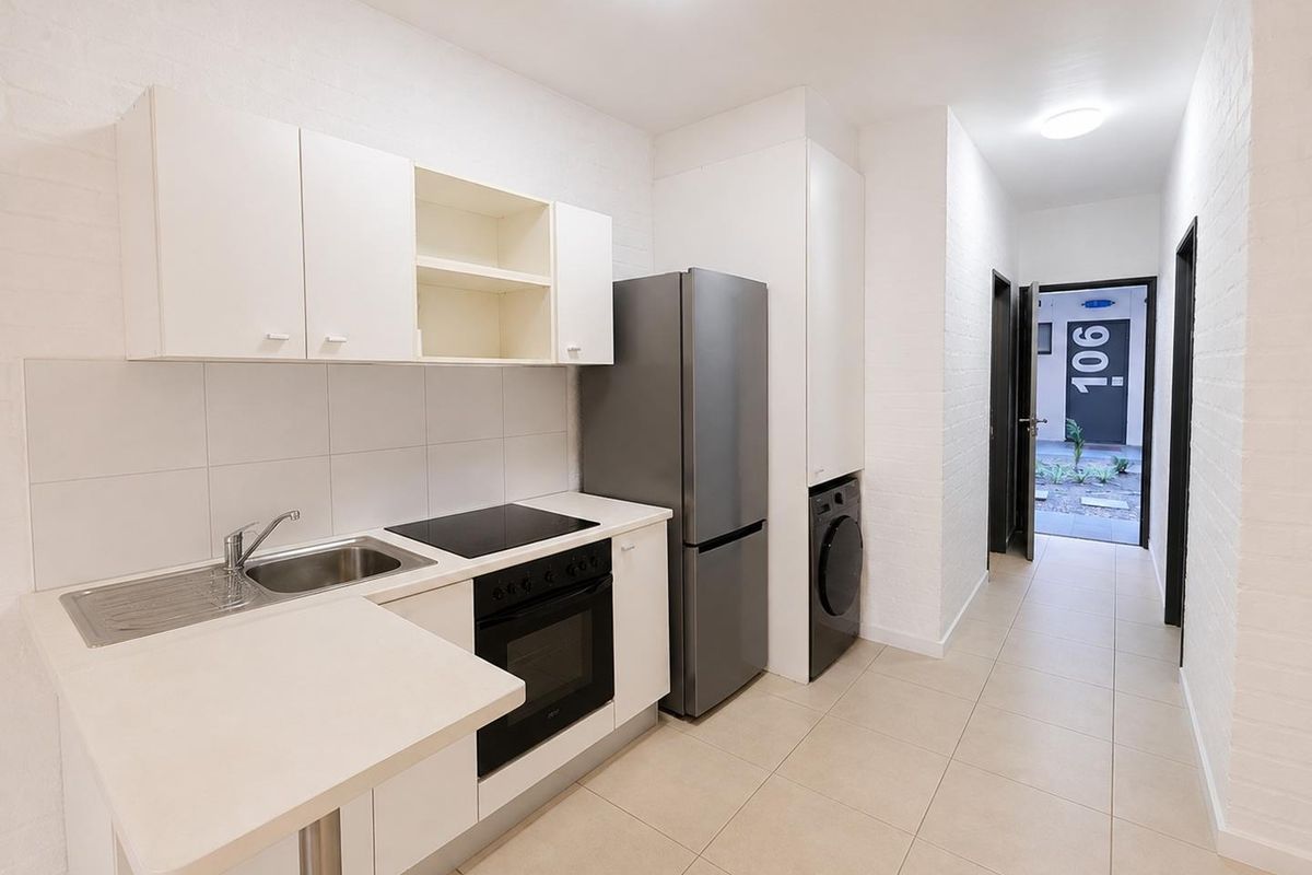 A clean, modern kitchen featuring white cabinetry, built-in oven and cooktop, tiled backsplash, and integrated laundry space, leading directly to the apartment entrance.