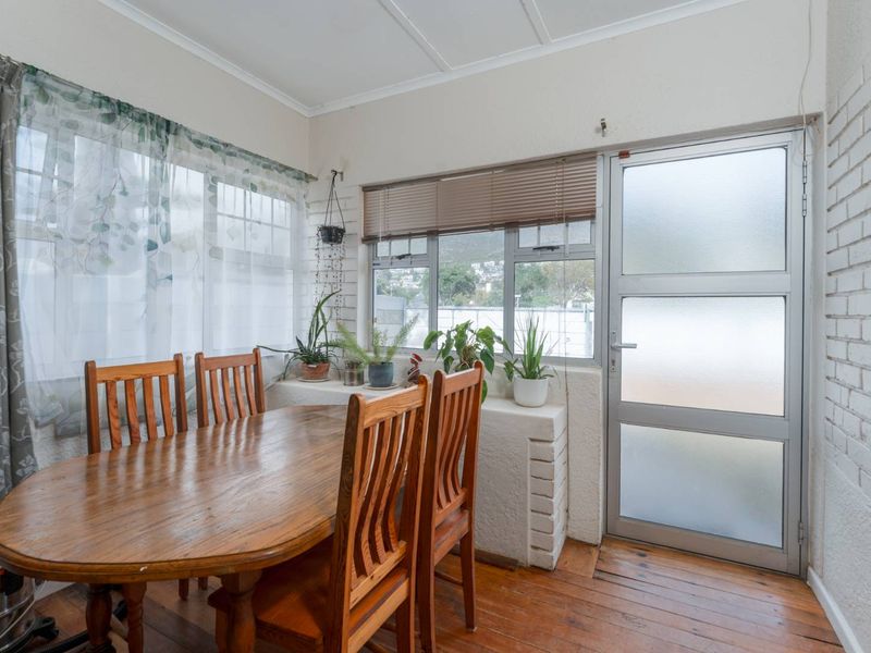 A dining area featuring timber flooring, painted brick walls, and natural light through aluminium-framed windows with a frosted glass exterior door.