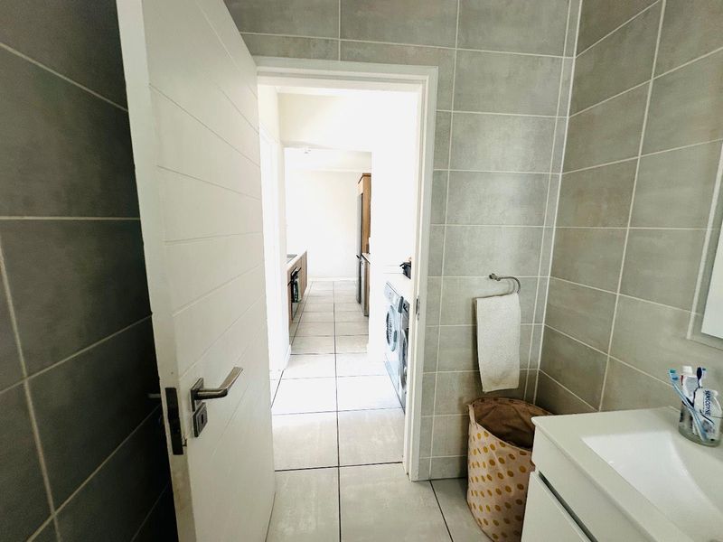 A view from a tiled bathroom looking through a doorway into an adjacent laundry and kitchen area with neutral finishes.
