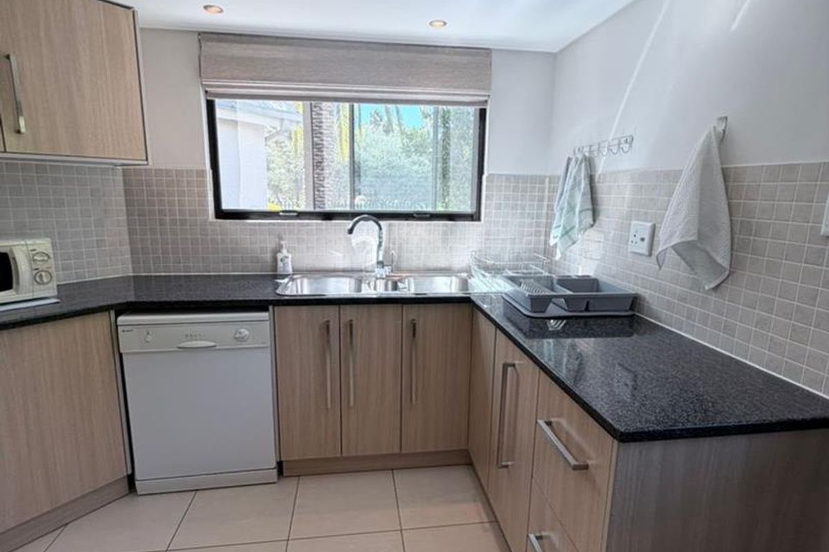 A functional kitchen featuring light wood-grain cabinetry, dark granite countertops, tiled splashbacks, and a double-basin sink with a window view.