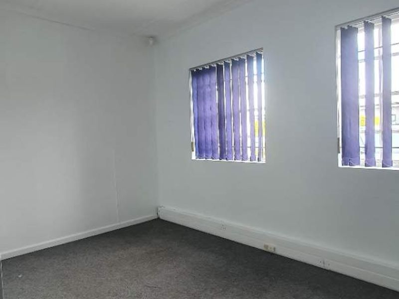 An empty office space featuring grey carpet flooring, white walls, vertical blinds, and security bars on the windows.