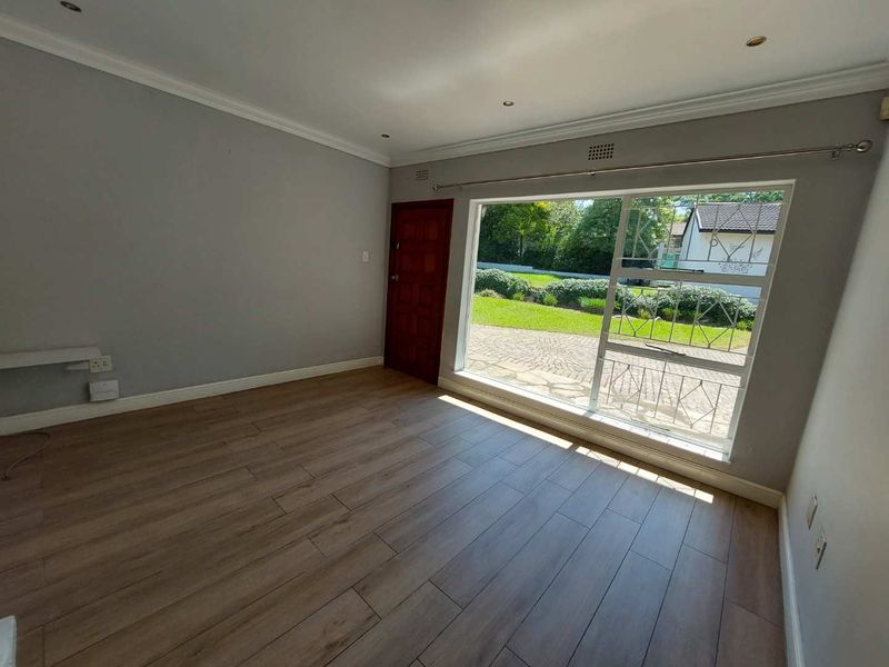 A bright, empty living room featuring modern laminate flooring, recessed downlights, and a large window with security bars overlooking the front garden.