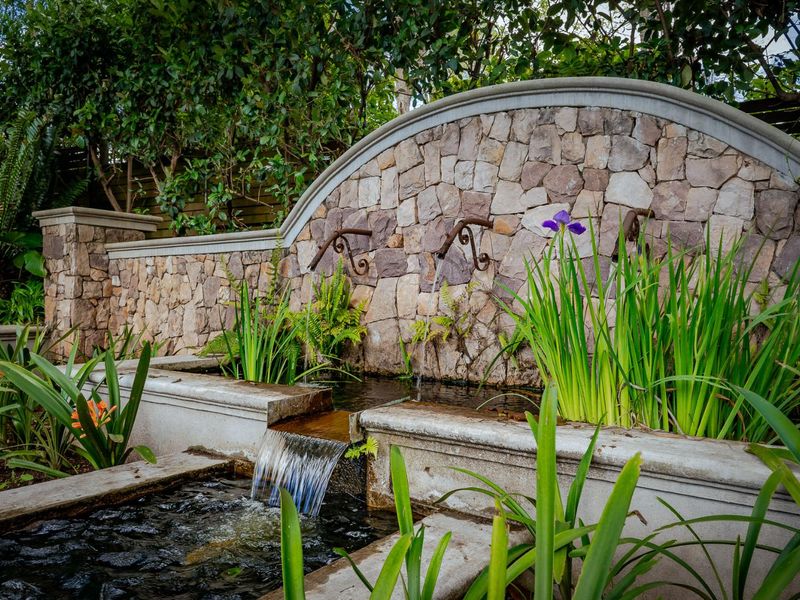 A beautifully landscaped garden area featuring a stone-clad water feature with cascading spouts and a concrete pond basin.