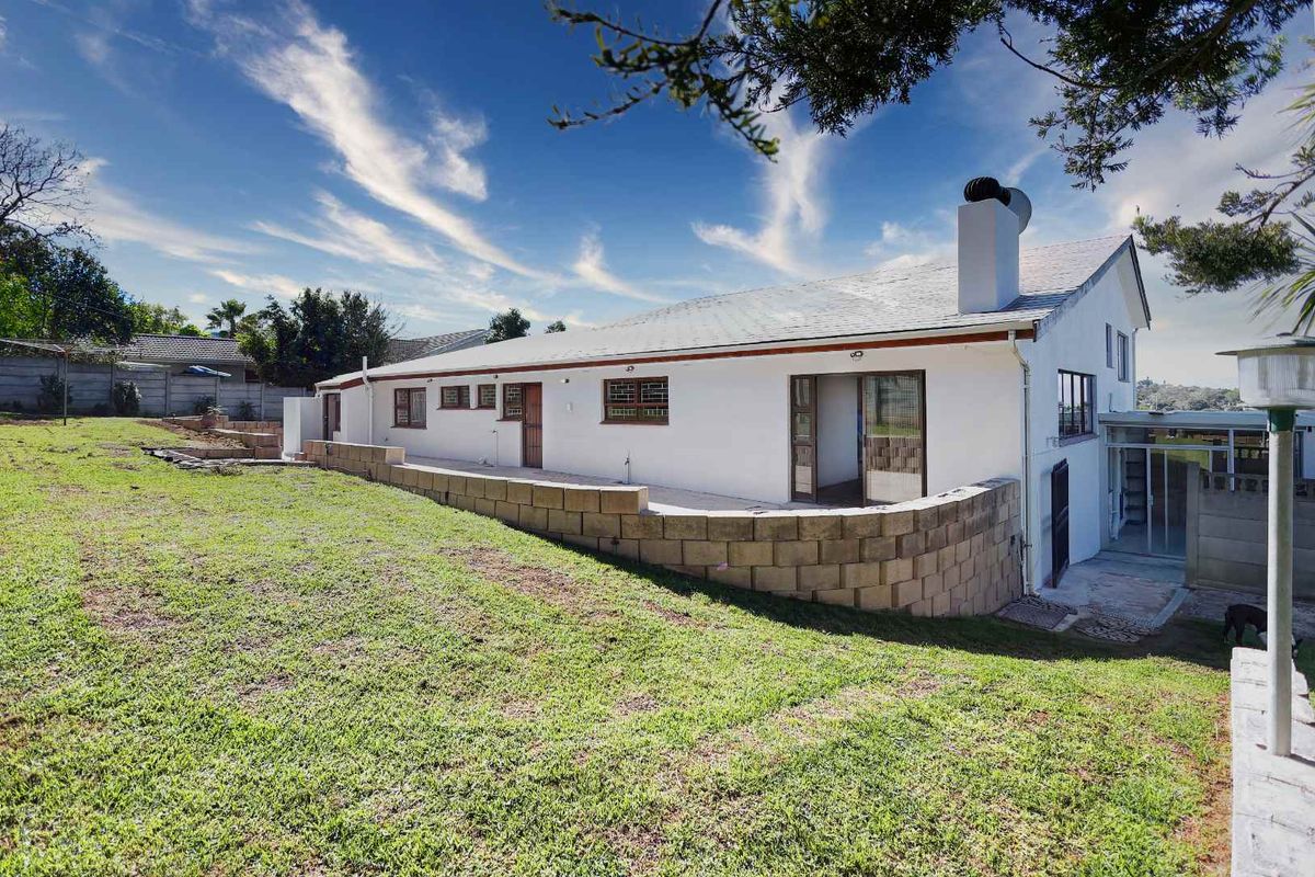 Rear view of the property featuring a white facade, a prominent chimney, a concrete retaining wall, and a spacious lawn area.
