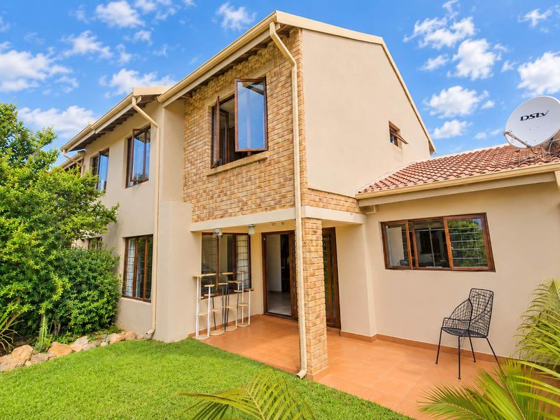 Rear view of a double-story townhouse featuring a tiled patio, brick and plaster facade, and well-maintained garden area.