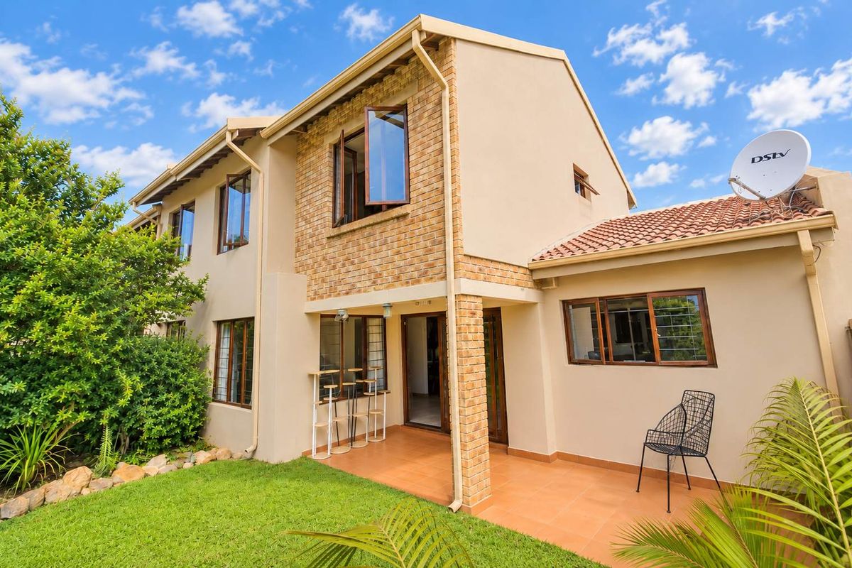 Rear view of a double-story townhouse featuring a tiled patio, brick and plaster facade, and well-maintained garden area.