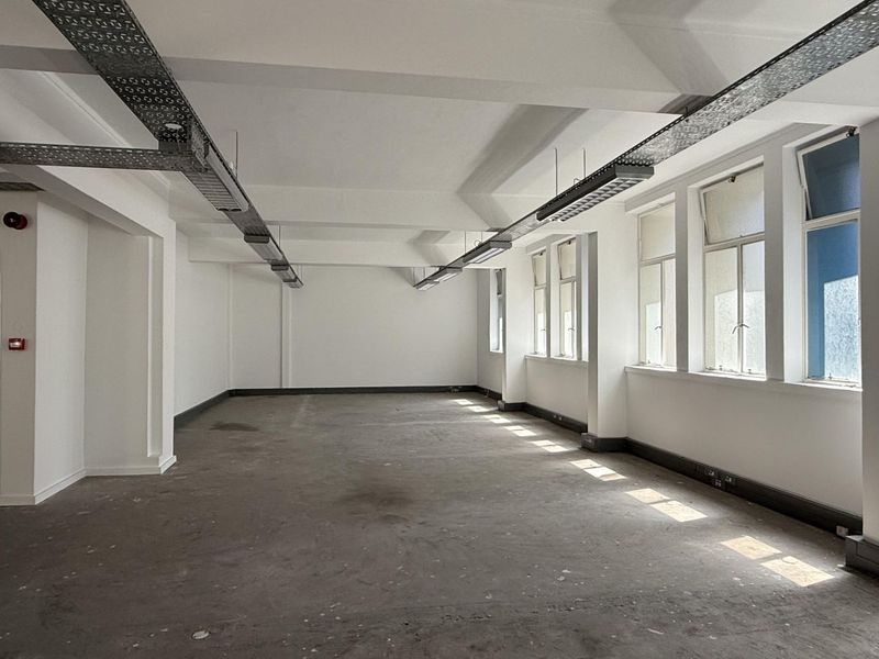 An open-plan office space featuring exposed industrial cable trays, large windows providing natural light, and durable concrete flooring.