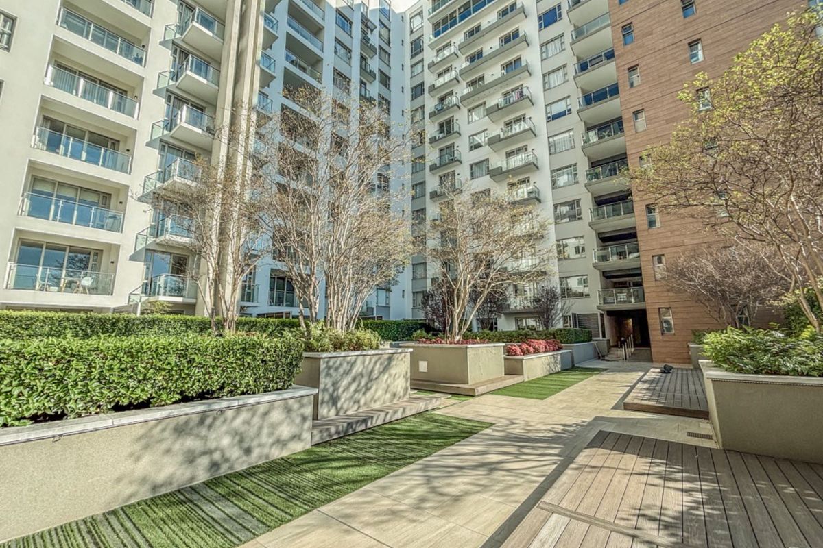 A well-maintained outdoor courtyard area featuring landscaped garden beds, paved walkways, and timber decking, surrounded by the modern apartment building facade.