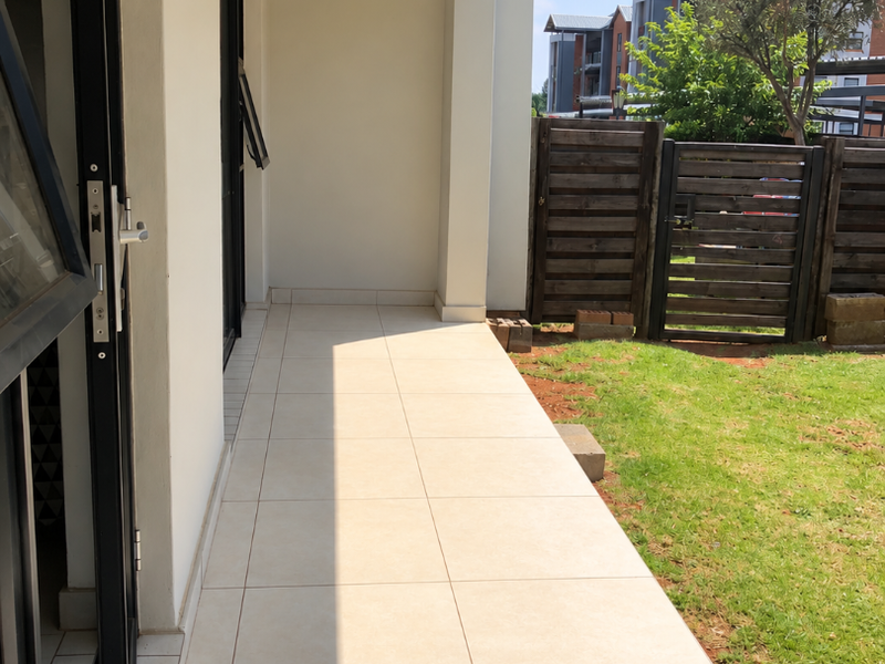 A clean, covered patio area featuring light-colored floor tiles, a ceiling light, and direct access to a private garden space with timber fencing.