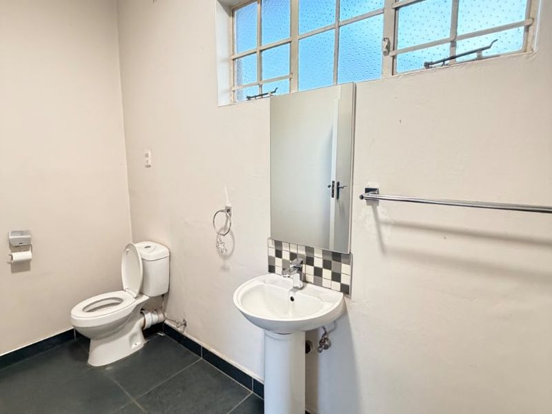 A functional bathroom featuring a pedestal basin, toilet, and dark tiled flooring, illuminated by a large steel-framed window.