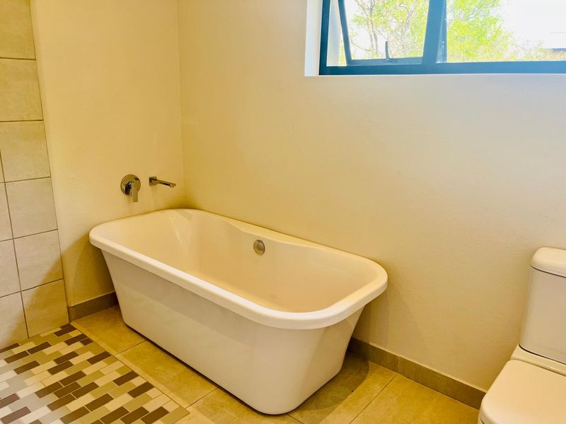 A clean, minimalist bathroom featuring a modern freestanding bathtub, wall-mounted fixtures, and neutral floor tiling with a top-hung window for natural light.