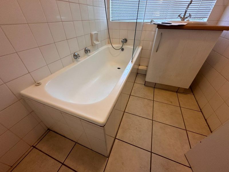A functional bathroom featuring a built-in bathtub with a glass screen, tiled surfaces, and a vanity cabinet, offering potential for cosmetic updates.