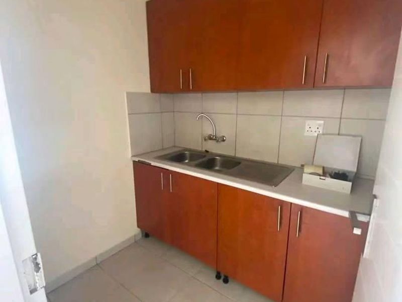 A compact kitchen area featuring wooden cabinetry, a stainless steel sink, tiled backsplash, and neutral floor tiles.