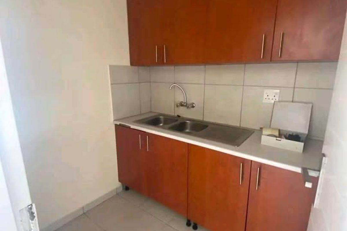 A compact kitchen area featuring wooden cabinetry, a stainless steel sink, tiled backsplash, and neutral floor tiles.