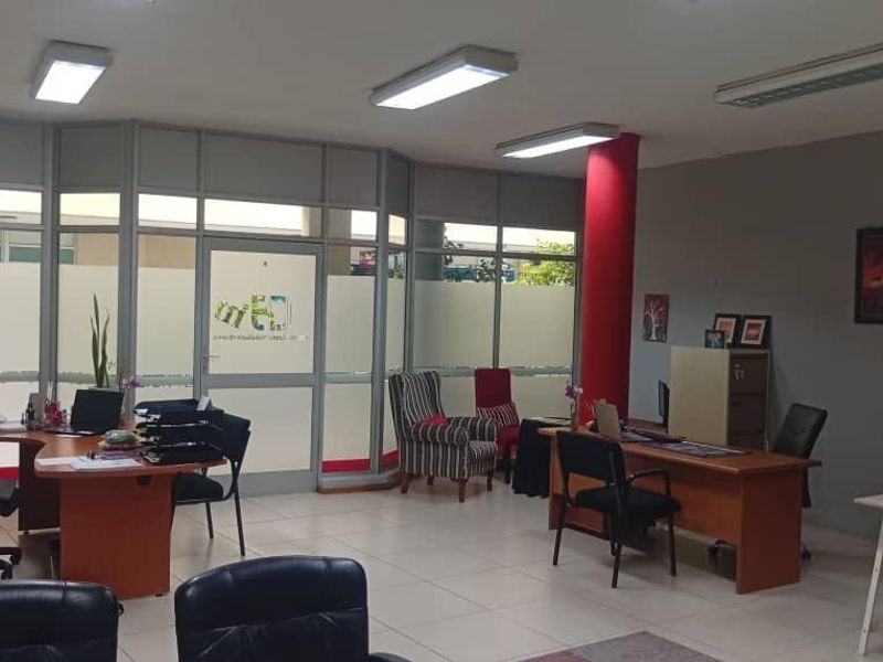 Open-plan office space featuring tiled flooring, aluminium-framed glass partitions, and integrated fluorescent lighting, providing a functional professional work environment.