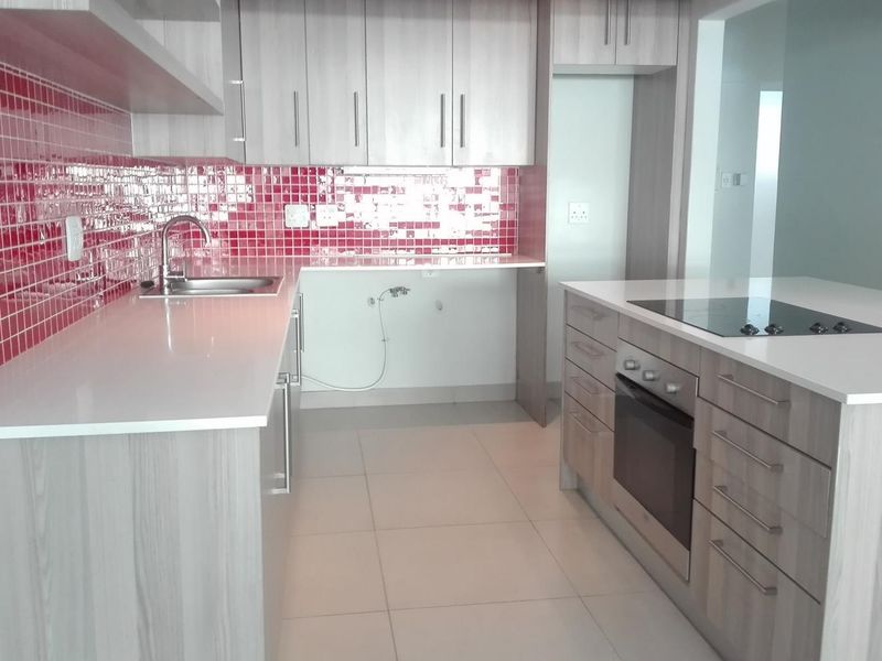 Modern kitchen featuring light wood-grain cabinetry, white quartz countertops, a striking red tiled backsplash, and a central island with a built-in oven and cooktop.