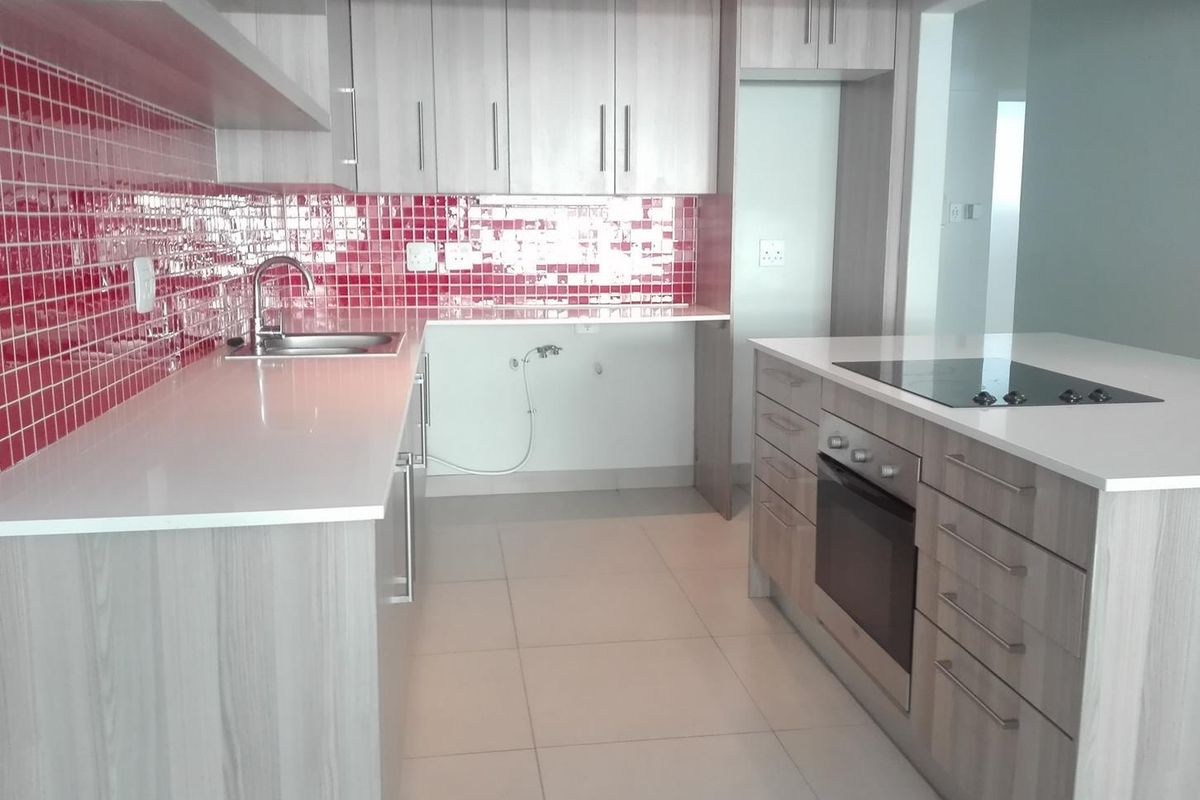 Modern kitchen featuring light wood-grain cabinetry, white quartz countertops, a striking red tiled backsplash, and a central island with a built-in oven and cooktop.