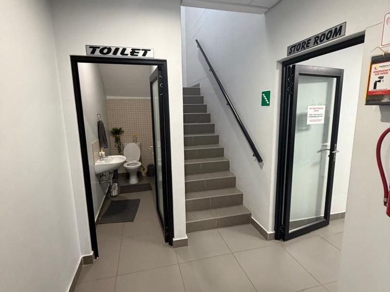 A clean, functional commercial hallway featuring tiled flooring, a central staircase with a handrail, and access to a restroom and storage room.